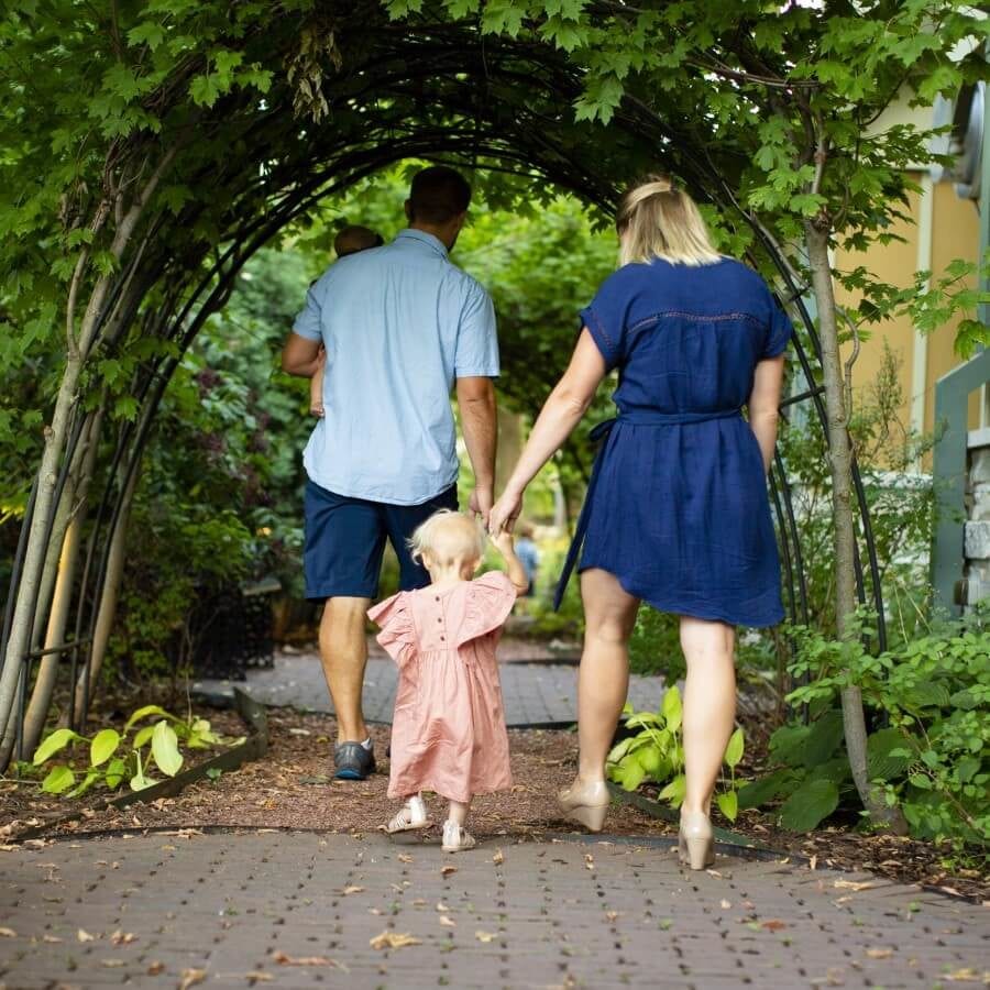 Little girl in pink dress holds her mother and father's hands as they walk under a garden archway.