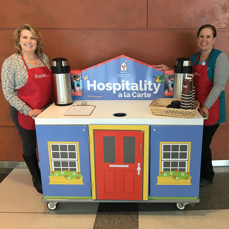 Two women stand on either side of a cart that looks like a small house that says "Hospitality a la Carte" with two coffee carafes on top of the cart.