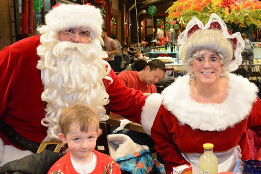 A young boy sits in front of a man and woman dressed as Santa Claus and Mrs. Claus.