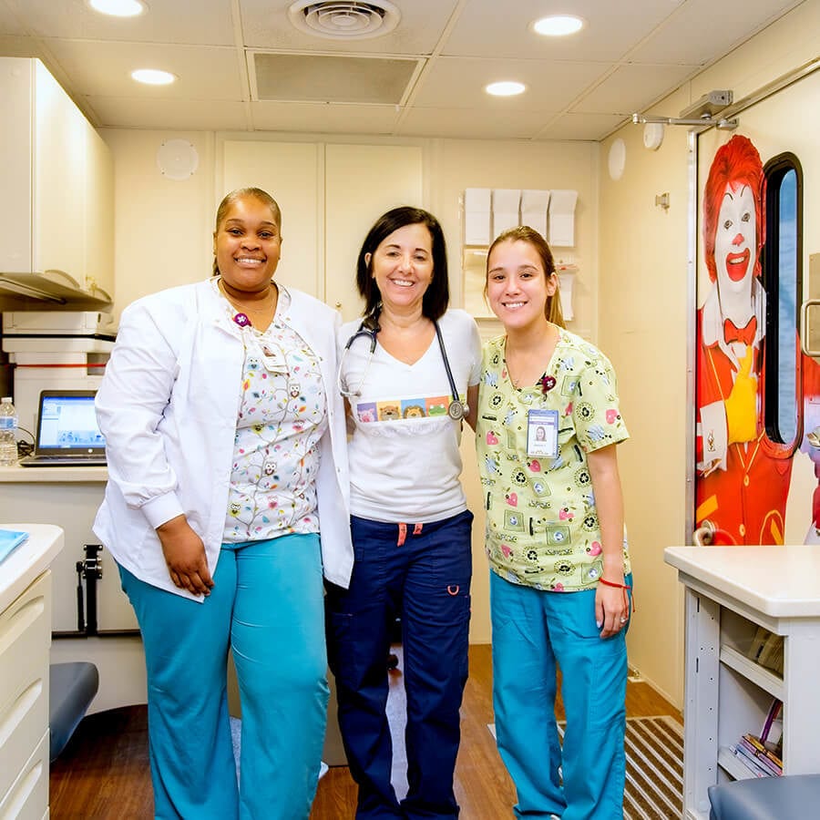 Three woman wearing hospital scrubs stand inside a small medical exam room.