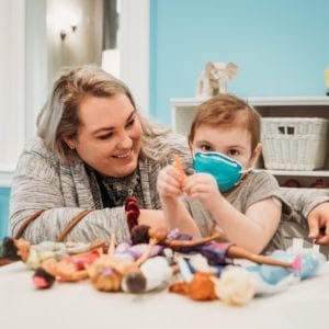Mother sits next to her daughter, who wears a medical mask, at a table full of toys