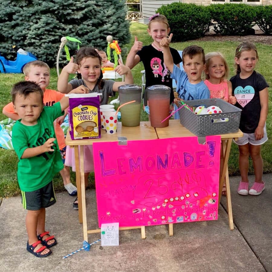 Several kids stand around a lemonade stand and give the thumbs up sign,