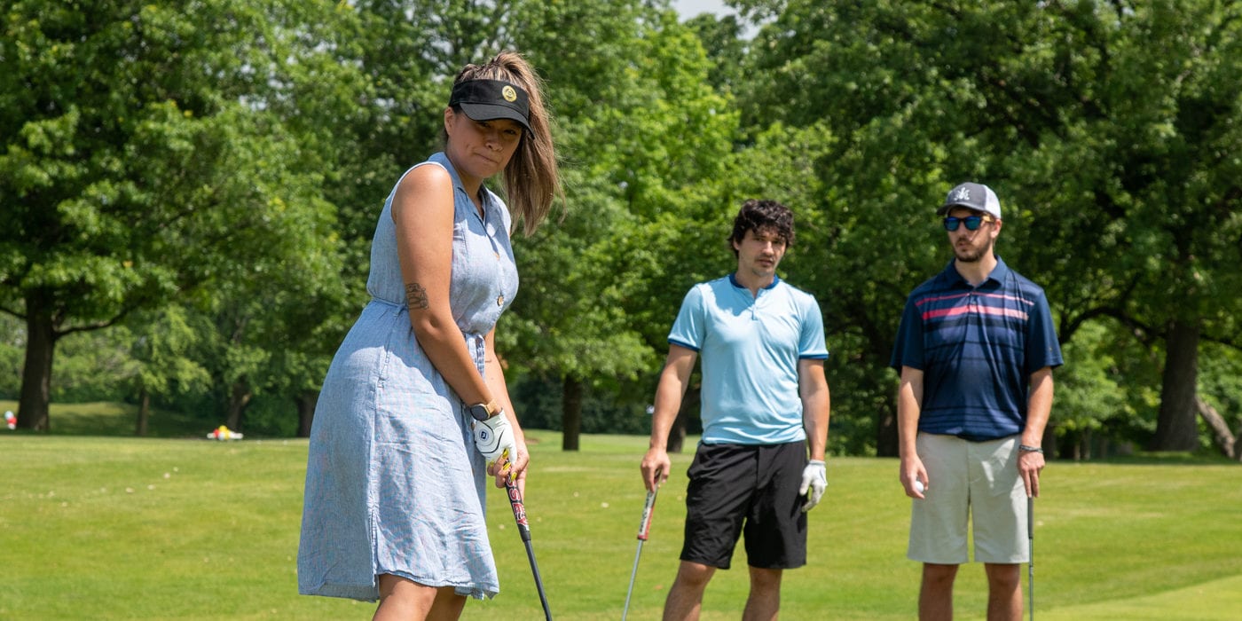 A white woman gets ready to put a golf ball white two men stand in the background.