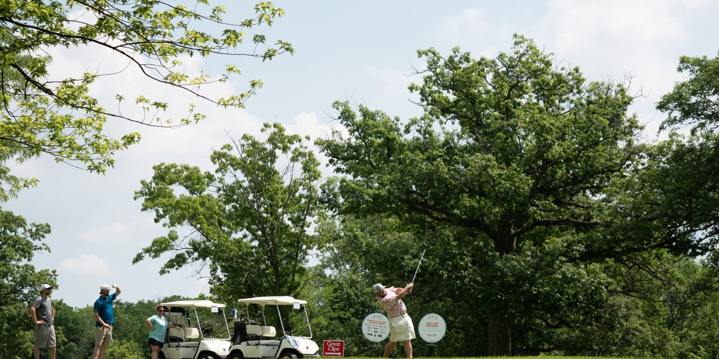 A white man swings a golf club with a golf cart behind him.