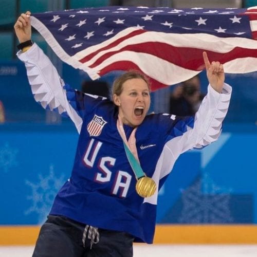 Female hockey player in gear with an Olympic gold medalwaves an American flag