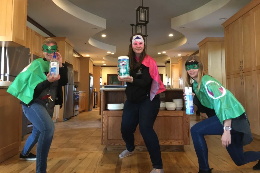 Three women pose in a kitchen with green masks and capes holding bottles of sanitizing wipes and Lysol spray.