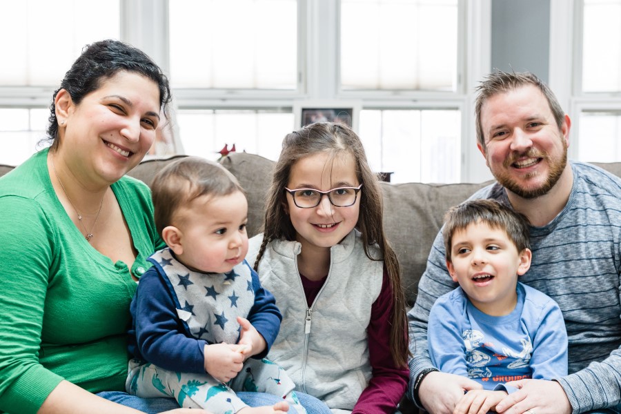 A mom and dad sit on a couch with a baby, a young boy, and a 10-year-old girl