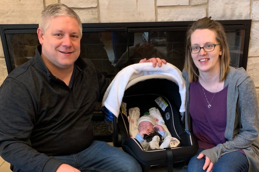 A man and woman with a baby in a carseat between them sit in front of a fireplace.