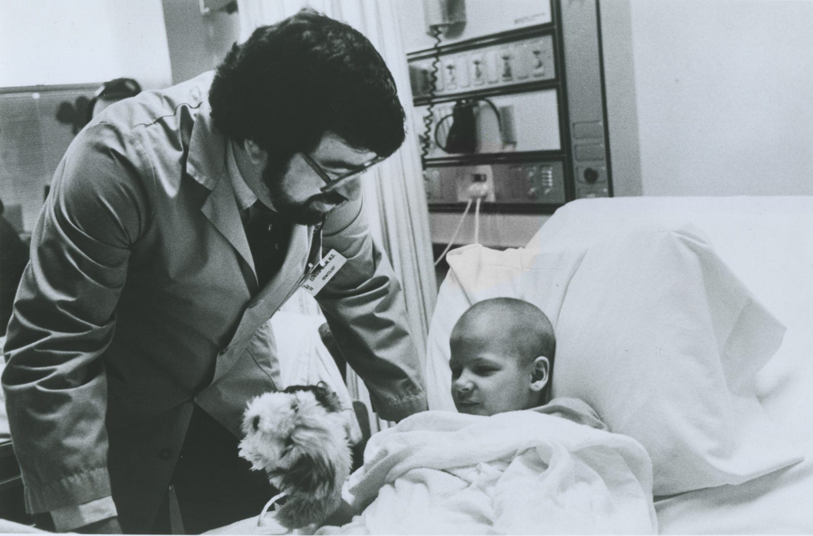 Black and white photo from the 1970s of a doctor with a black beard leaning over a child patient in a hospital bed holding a stuffed animal.