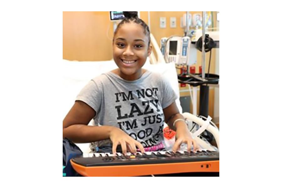 A Black teen girl sits in a hosital bed, smiling and playing a keyboard in her lap.