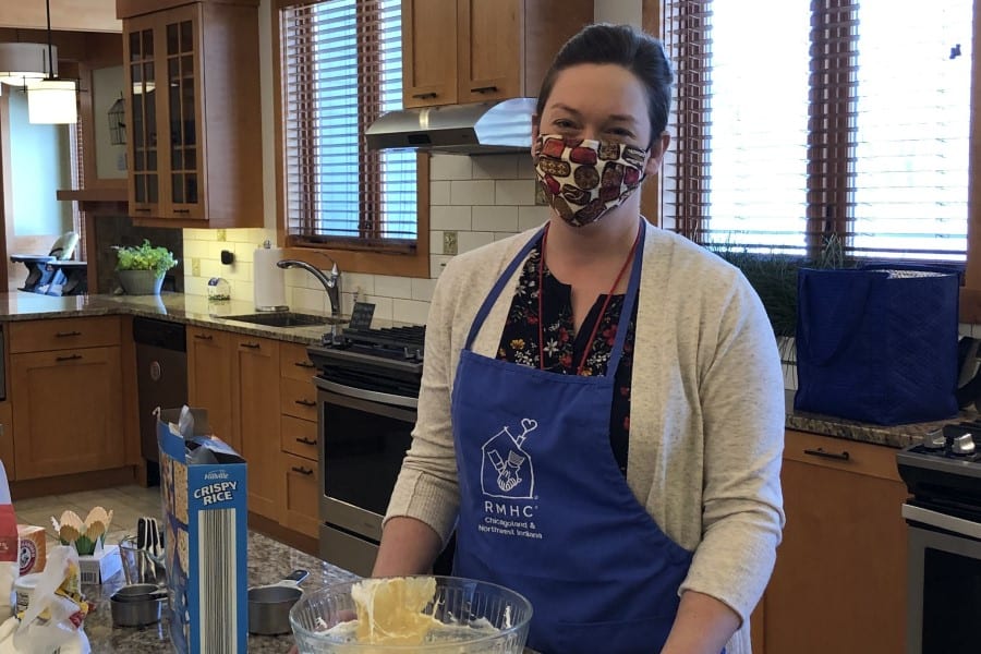 A woman in a fabric mask and RMHC apron stands in a kitchen cooking rice krispie treats.