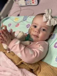 A baby girl with a pink onesie claps her hands on a hospital bed.