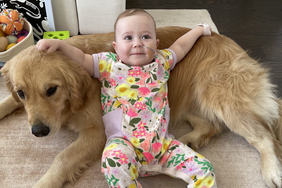 A baby in a flower onesie and a medical tube coming from her nose lounges against a golden retriever.